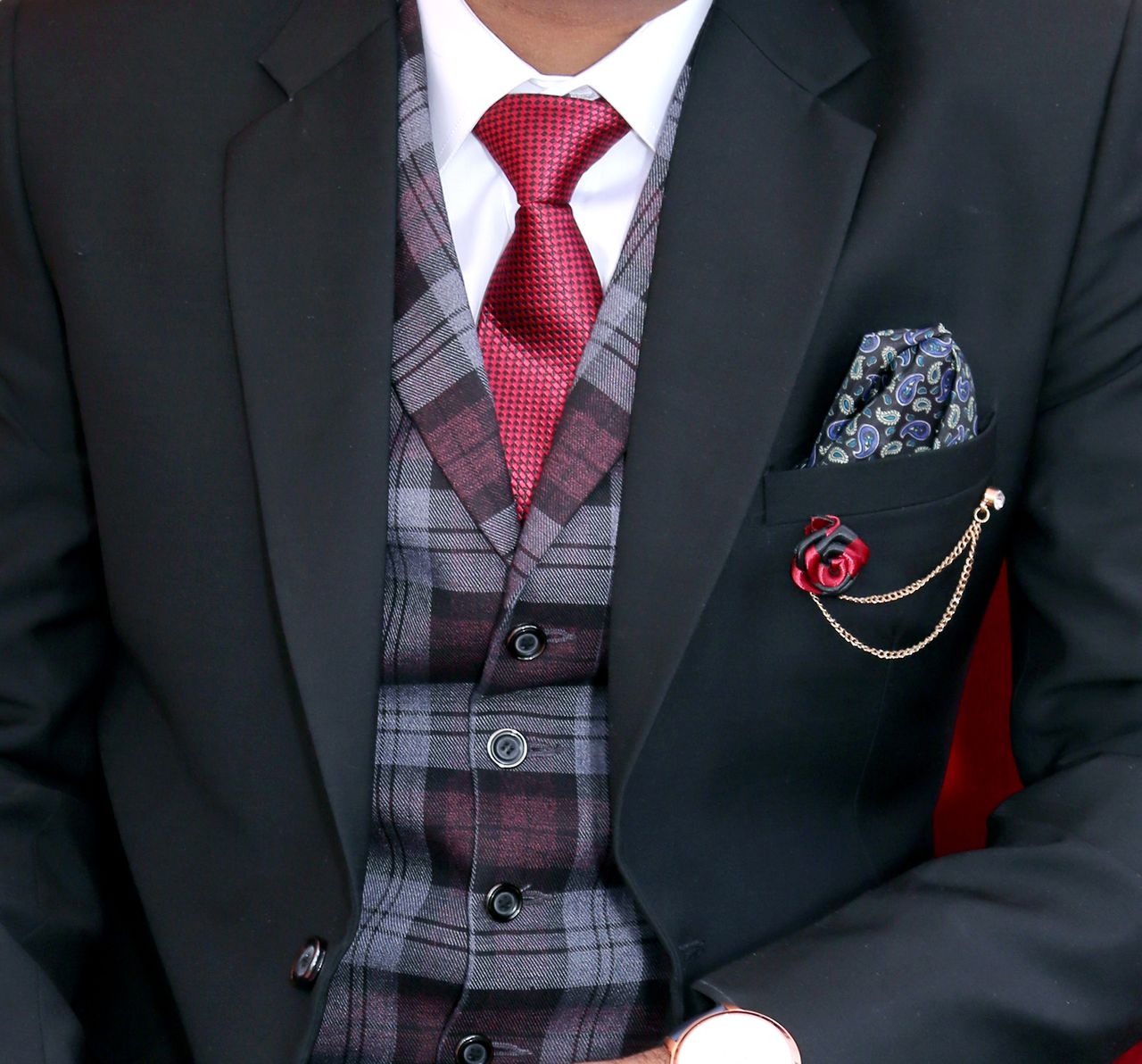 services-01 Close-up of a man in an elegant black suit with a red tie and plaid vest, showcasing formal menswear.